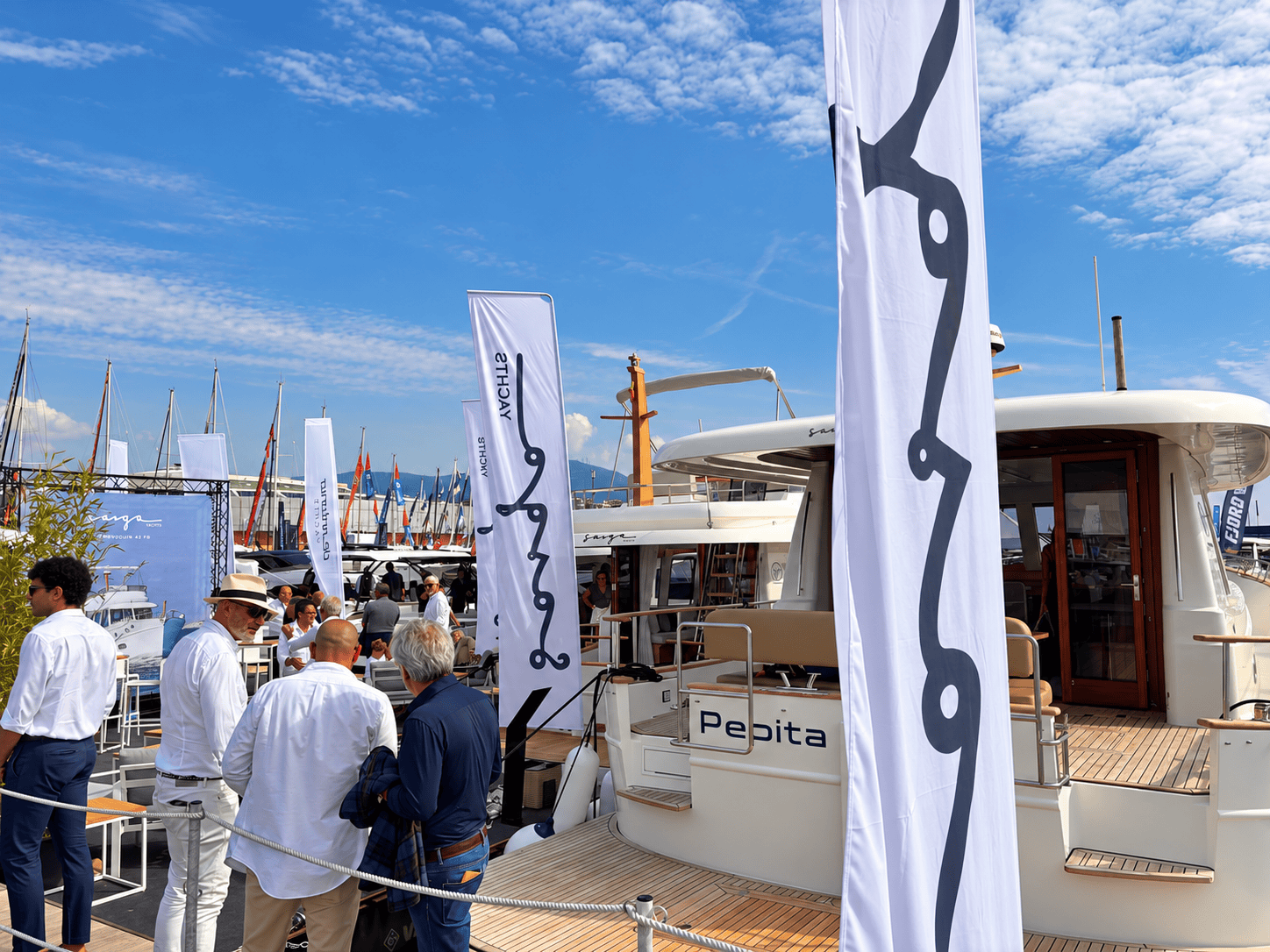 Banderas de Sasga Yachts frente a la popa del Menorquín Pepita, con visitantes reunidos en el pantalán.