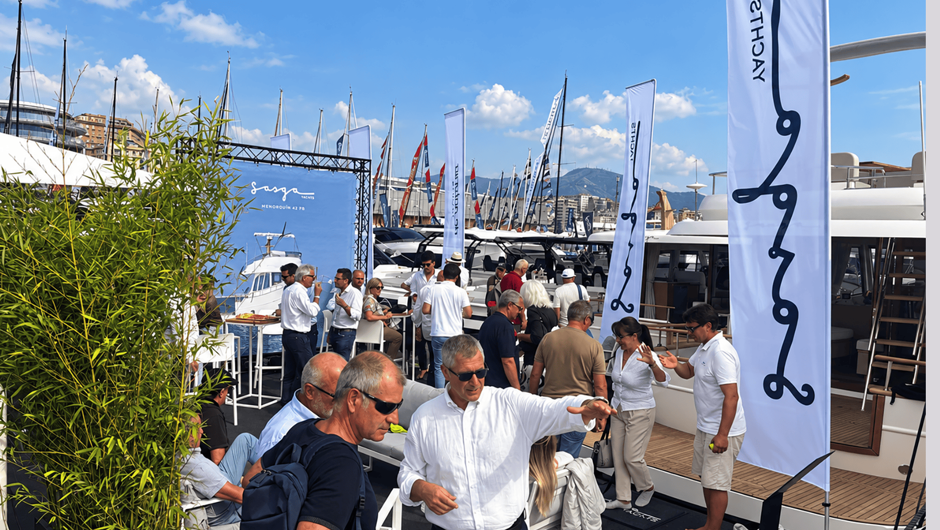 Visitantes conversando en el stand de Sasga Yachts, entre banderas de la marca y barcos amarrados al pantalán. Salón Náutico de Génova 2026